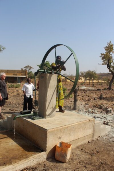 Une femme portant une robe de couleur jaune et verte puise de l'eau en actionnant avec sa main la roue du puits.