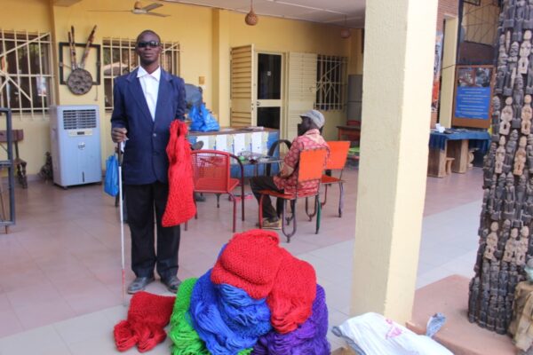 Dans un jardin à Ouagadougou, Casimir debout tient sa canne blanche dans sa main droite et, dans la gauche, des filets rouges. À ses pieds, un gros de tas de filets rouges, verts, bleus et mauves. Derrière lui, une table avec un homme assis de dos.