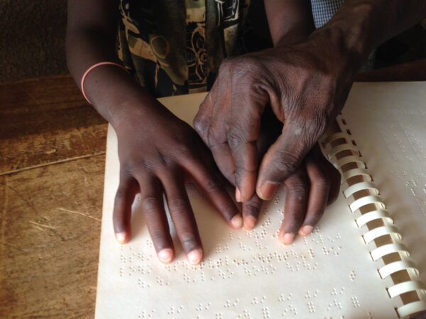 Au-dessus d'un livre en braille, la main du professeur guide les mains d'un enfant en train de lire.