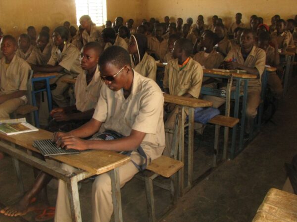 Une salle pleine de lycéens en uniforme. Au premier plan, Sidiki est en train d'écrire le braille sur sa tablette avec une sacoche sur les genoux.