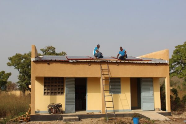 Deux ouvriers sont sur le toit d'un bâtiment en train de poser les panneaux solaires. Une échelle est posée contre le bâtiment comportant deux portes grises. En arrière-plan, des arbres et le ciel bleu.