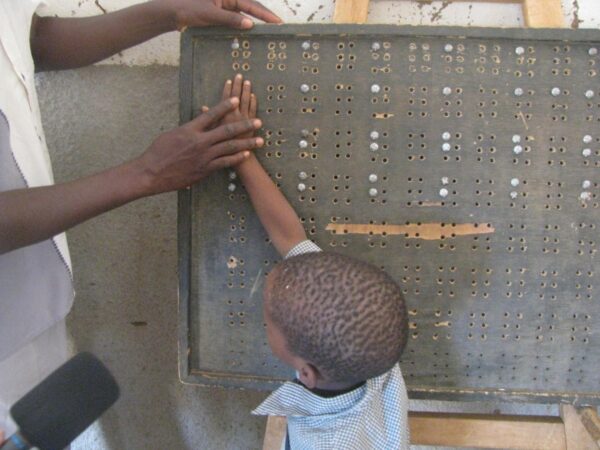 Un panneau en bois est posé verticalement sur un support. Le panneau est couvert des cases comportant les six trous composant la matrice du braille les points sont constitués par des vis de récupération. Un petit garçon a la main posée sur le panneau. Un adulte est en train de guider sa main.