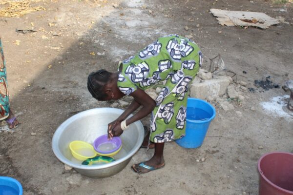 Dans une cour terreuse, une jeune fille vêtue d'une robe verte avec des motifs gris, blancs et bleus fait sa vaisselle penchée au-dessus d'une bassine en fer blanc. Derrière elle, un sceau bleu en plastique contenant de l'eau.