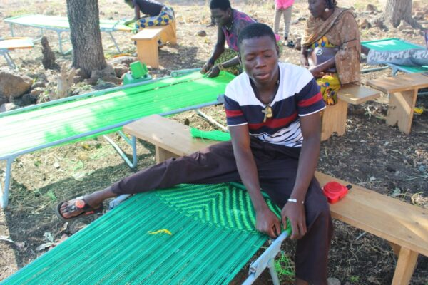 Au premier plan, un jeune homme aveugle assis sur un banc, tisse un sommier. Au second plan, deux autres tisserandes et une autre femme assise sous les arbres.
