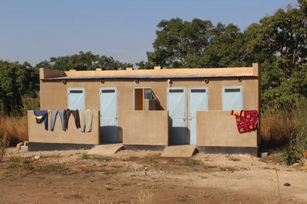 Un bâtiment comportant six portes abrite les douches et les latrines. Des vêtements sèchent au soleil. En arrière-plan, des arbres et de la végétation.