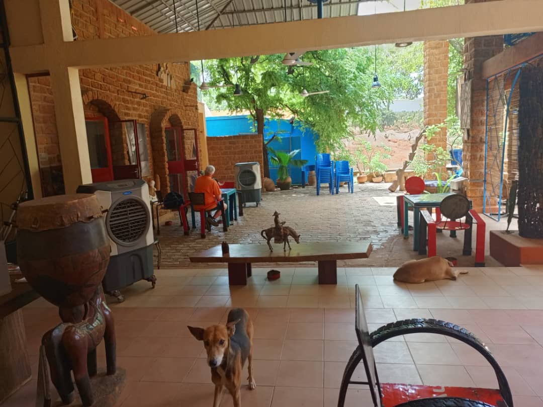 Be My AI: La photo montre une cour intérieure avec un toit en tôle ondulée. À gauche, il y a un mur en briques avec des portes et des fenêtres rouges. Un homme portant un t-shirt orange est assis à une table rouge et verte, probablement en train de travailler ou de lire. Il y a plusieurs objets et meubles dans la cour, y compris des ventilateurs portables, des chaises empilées bleues, et des tables avec des chaises assorties. Au premier plan, il y a une sculpture en bois et un tambour. Deux chiens sont visibles, l'un debout au centre de la photo et l'autre couché sur le sol à droite.
Des plantes en pot et un grand arbre ajoutent une touche de verdure à l'espace. L'ambiance générale est détendue et ombragée, avec des éléments de décoration rustiques et artistiques.