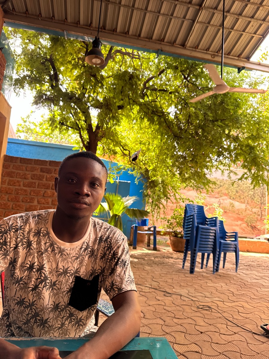 Be My AI: La photo montre un jeune homme assis à une table en extérieur. Il porte un t-shirt blanc avec des motifs de palmiers noirs et une poche noire sur le côté gauche de sa poitrine. Il a la peau foncée et regarde directement vers la caméra avec une expression neutre.En arrière-plan, on peut voir un arbre avec un feuillage dense, ce qui donne une impression de fraîcheur et d'ombre. Il y a également un mur en briques rouges et un mur peint en bleu. À droite de l'image, il y a plusieurs chaises en plastique bleu empilées les unes sur les autres. Le sol est pavé de carreaux de couleur beige.
Le lieu semble être une terrasse couverte, car on peut voir un toit en tôle au-dessus de l'homme, avec une lampe suspendue et un ventilateur de plafond.